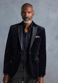 Blazer negro de terciopelo con solapas de satén, combinado con una camisa oscura. Incluye un pañuelo de bolsillo con un patrón. Ajuste limpio y entallado con bolsillos laterales.