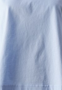 Ljusblå bomulls t-shirt med en slät textur, rundad halsringning och ett subtilt, något rynkat utseende. Sömmen är synlig vid fållen.