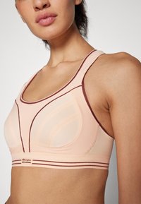 Ferskenfarget sports-bh med mesh- og stoffpaneler. Har scoop-neck, teksturerte detaljer og vinrødt kantbånd. Merkevaren leser "Champion Shock Absorber."