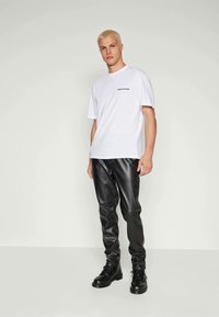 Weißes Baumwoll-T-Shirt mit schwarzem Logo auf der linken Brust, kombiniert mit schwarzen Lederhosen und schwarzen Schnürstiefeln.