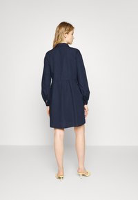 Vestido camisola azul marinho com mangas longas, cintura ajustada e saia rodada. Apresenta um colarinho e punhos abotoados. Usado com mules amarelas de salto.