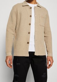 Cárdigan de punto beige con un patrón texturizado, parte delantera abotonada y cuello. Cuenta con tres botones en la parte delantera y un bolsillo de parche en el lado izquierdo.