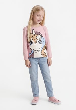 Smaidīga meitene ar gariem blondiem matiem valkā rozā garo piedurkņu kreklu ar vienradža grafiku, gaiši zilas džinsas un rozā sporta apavus, stāv atvieglināti.