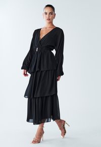 Langärmliges schwarzes Kleid mit tiefem V-Ausschnitt, tailliertem Gürtel, mehrlagigem Rock und fließendem Stoff. Kombiniert mit goldenen, knappgeschnürten High-Heel-Sandalen.