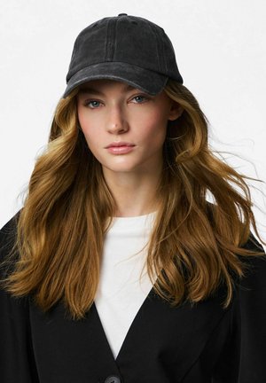 Mujer joven con cabello largo y ondulado de color castaño claro, que lleva una gorra negra, una camiseta blanca y una chaqueta negra, mirando directamente a la cámara.