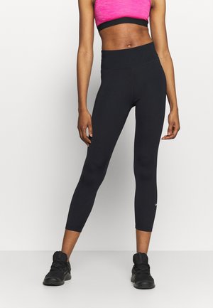 Persona atlética con leggings negros cortos, sujetador deportivo rosa y zapatillas negras, de pie con las piernas separadas sobre un fondo blanco.