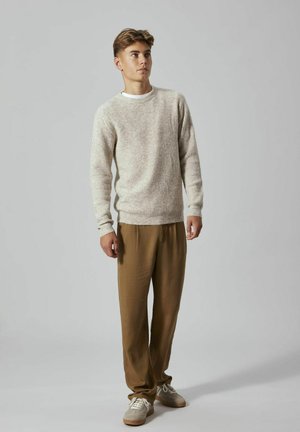 Maglione a maglia beige con texture a coste, abbinato a pantaloni larghi marroni e sneakers grigie con lacci bianchi. Design semplice e moderno.