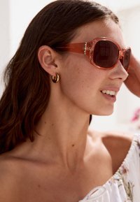 Oversized solglasögon med en genomskinlig orange ram och mörka glas. Har dekorativa detaljer på skalmarna. Enkla öronringare.