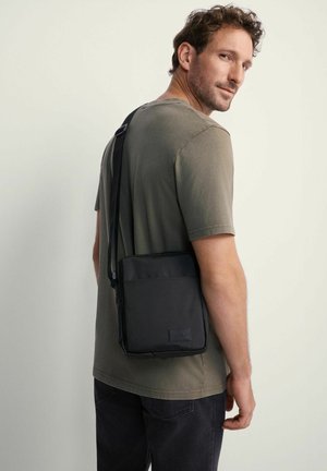 Czarna torba na ramię wykonana z trwałego materiału, o eleganckim prostokątnym kształcie, z regulowanym paskiem oraz przednią kieszenią z metką marki.