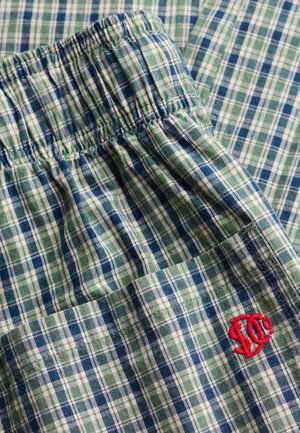 Grønt, blåt og hvidt ternet stof med elastisk linning og en rød broderet logo på en lomme.