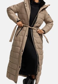 Persona que lleva un abrigo largo de tela acolchada beige con capucha, atando el cinturón sobre un vestido negro de canalé y botas negras de tobillo.