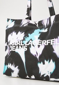 Veľká taška z látky, s čiernym základom a bielymi a svetlomodrými kvetinovými vzormi. Text znie "KARL LAGERFELD JEANS."