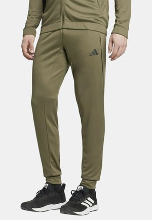 Pantaloni sportivi verde oliva con design sagomato, dotati di tasche laterali e logo Adidas nero sulla coscia sinistra. Tessuto liscio.