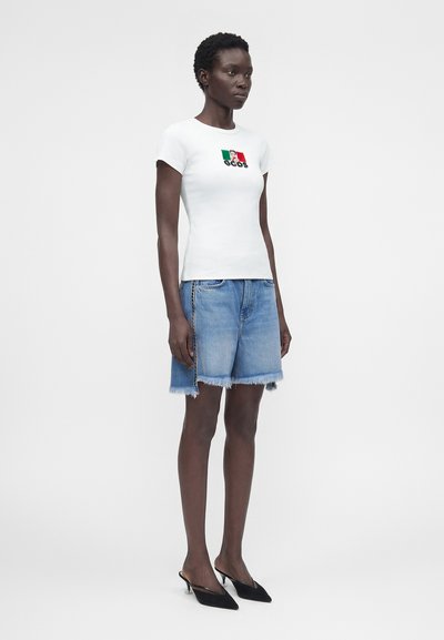 Γυναίκα με λευκό εφαρμοστό μπλουζάκι με τη σημαία και το λογότυπο της Ιταλίας, φθαρμένα μπλε τζιν σορτς και μαύρα μυτερά mules με kitten heel, στέκεται πλαγίως.