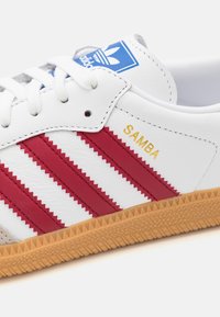 White leather sneaker with red zigzag stripes and gold "SAMBA" text. Blue Adidas logo on tongue, brown rubber sole with patterned texture.