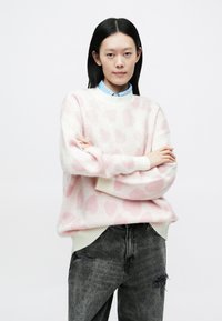 Gaiši rozā džemperis ar mīkstu tekstūru, tam raksturīgs tumšāku rozā plankumu raksts, ribains apakšmala un brīva piegriezuma siluets. Uzvilkts kopā ar pelēkām izdilušām džinsām.