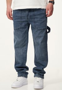 Dunkelblaue Denim Cargo-Hosen mit lässiger Passform. Verfügt über mehrere Taschen, sichtbare Nähte und ein seitliches Riemendetail. Kombiniert mit weißen Sneakers.