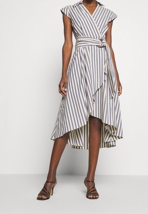 Femme portant une robe rayée sans manches, dégradée, avec une ceinture à la taille et des sandales noires à talons hauts et brides.