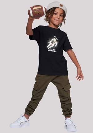 Black t-shirt with "FUTURE ASTRONAUT" graphic, featuring an astronaut. Styled with cargo pants and white sneakers, holding a football.