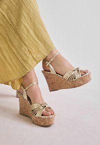 Zlaté wedge sandále s korkovou platformou, so skríženými remienkami a detailom uzla vpredu. Remienok okolo členku obsahuje pracku na nastavenie.