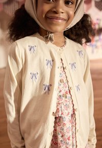 Cream cardigan with blue embroidered bows, button closure, and ribbed cuffs, layered over a floral dress with ruffle detailing and buttons.