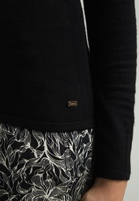 Pull en maille noire à coupe ajustée, avec une petite étiquette de marque dorée. Associé à un tissu à motif floral noir et blanc.