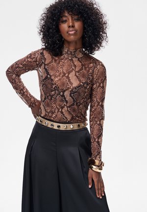 Kobieta o kręconych włosach ma na sobie brązowy golf w wężowy wzór, czarną spódnicę z ozdobnym paskiem i masywne złote bransoletki, pozując pewnie.