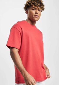 Ung mann med krøllete hår iført en ensfarget rød oversized t-skjorte, holder i kanten, mot en ensfarget lys bakgrunn.