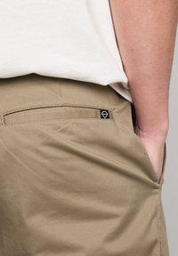 Khaki pants with a smooth texture, featuring a back pocket and a small black logo tag. The fabric is lightweight and durable.
