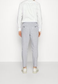 Man wearing light grey slim-fit trousers, white dress shirt, and off-white shoes standing against a plain background, back facing viewer.