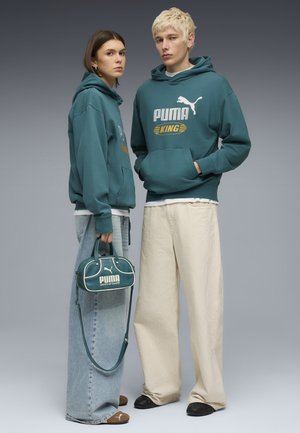 Twee modellen dragen tealkleurige Puma King hoodies; een houdt een tealkleurige Puma handtas vast en draagt brede jeans, de ander draagt losse crèmekleurige broek.