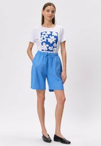 Biała koszulka z niebieskim kwiatowym nadrukiem, połączona z jasnoniebieskimi szortami z elastycznym pasem i czarnymi płaskimi butami. Prosty i casualowy styl.