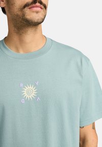 Hellblaues Baumwoll-T-Shirt mit einem Sonnenmotiv in Creme und Lavendel, umgeben von den Buchstaben "RVCA". Rundhalsausschnitt und kurze Ärmel.