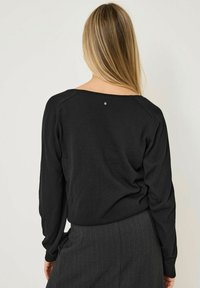 Kvinna med långt blont hår som bär en svart långärmad topp och mörkgrå randiga byxor, vänd bort från kameran mot en enfärgad bakgrund.