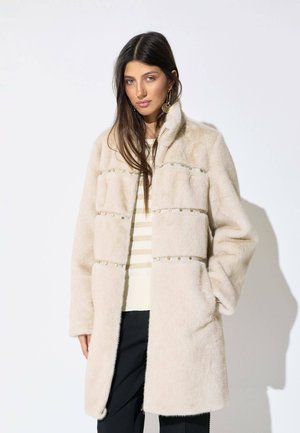 Mujer con cabello largo y oscuro, vestida con un abrigo beige peludo con tachuelas metálicas, camiseta de rayas y pantalones negros, de pie contra un fondo blanco.