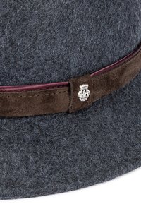 Grauer Hut mit breiter Krempe, akzentuiert durch ein dunkelbraunes Wildlederband und ein silbernes Emblem. Texturierte Oberfläche sichtbar.