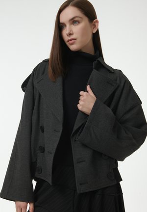 Chaqueta gris corta con mangas anchas, cierre de botones dobles y una tela texturizada. Llevada sobre un suéter negro de cuello alto.