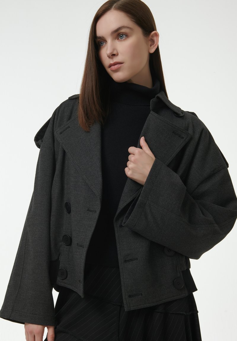 Grijze cropped jas met brede mouwen, dubbele knoopsluiting en een gestructureerde stof. Draagt over een zwarte coltrui.