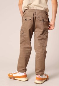 Bruine cargo-broeken met dubbele zijzakken, ritsen en knoopzakken achter. Gemaakt in combinatie met meerkleurige sneakers en witte sokken.