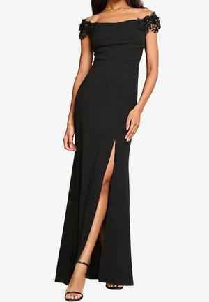 Femme portant une robe de soirée noire épaules dénudées avec des manches en dentelle florale perlée et une fente haute sur la cuisse, assortie à des talons noirs à bride à la cheville.