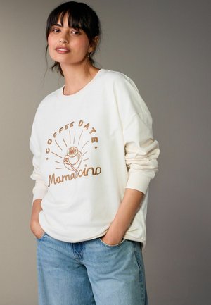 Žena s tmavými vlasmi v bielej mikine s nápisom a ilustráciou „COFFEE DATE Mamacino“, v svetlomodrých džínsach, ruky vreckách.