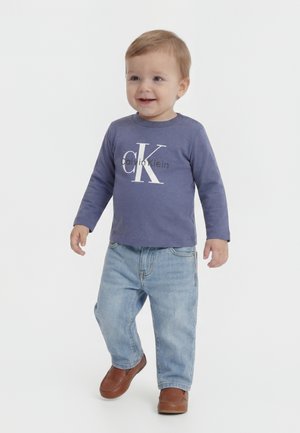 Kleinkind lächelt, trägt ein blaues langärmeliges Calvin Klein-Shirt, hellblaue Jeans und braune Schuhe, steht auf weißem Hintergrund.