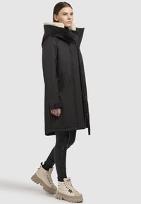 Schwarzer isolierter langer Mantel mit Kapuze und Lammfellfutter, ausgestattet mit einem Reißverschluss vorne und seitlichen Taschen, kombiniert mit beigefarbenen klobigen Stiefeln.