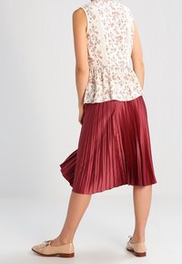 Ärmellose Bluse mit floralem Muster und Peplum-Saum, kombiniert mit einem plissierten burgunderfarbenen Rock und beigen Loafers mit Schleifendetail, die eine glatte Textur aufweisen.