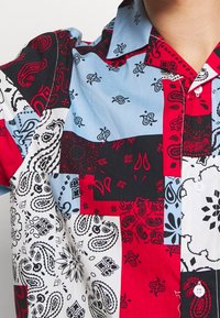 Camicia a maniche corte con un design a patchwork in rosso, blu, nero e bianco, con motivi paisley e floreali e chiusura a bottoni.