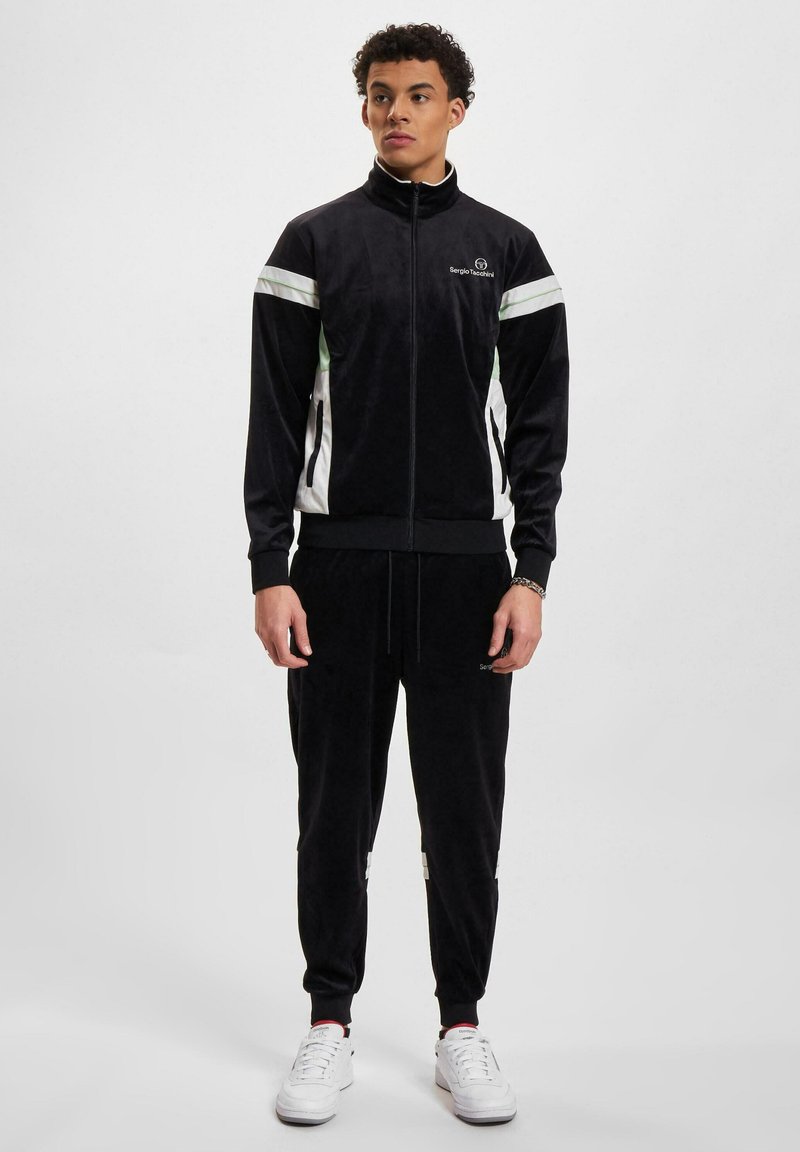 Ung mann står iført svart Sergio Tacchini treningsdrakt med hvite og grønne detaljer, sammen med hvite joggesko foran en ensfarget bakgrunn.