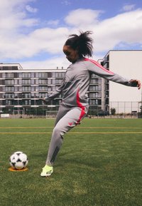 Šedý sportovní komplet s červenými akcenty, pruhované detaily na rukávech a kalhotách; zelené fotbalové kopačky; černobílý fotbal na travnatém hřišti.