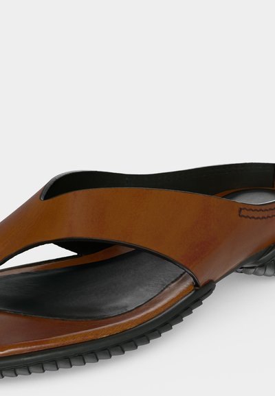 Καφέ δερμάτινο σανδάλι τύπου slip-on με μαύρο μαξιλαρωμένο πέλμα και μαύρη ελαστική σόλα με υφή, σε γωνιακή όψη σε λευκό φόντο.