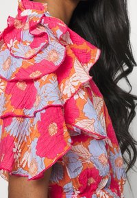 Bunt gemusterte Bluse mit pinken und blauen Blumen auf orangefarbenem Hintergrund, verziert mit Rüschendetails und einem strukturierten Stoff.