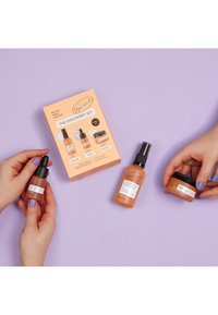 Entdeckungsset in einer orangefarbenen Box. Enthält drei braune Glasflaschen: eine Tropfflasche, einen Spray und ein Cremeglas mit schwarzen Akzenten. Lavendelfarbener Hintergrund.